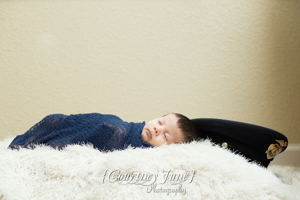 newborn photographer photographing a newborn swaddled in a navy blanket on a furry rug with a police officer's hat